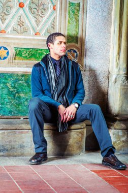 Young man, sitting against wall in winter, thinking, wearing in woolen jacket, tie, long scarf, black pants, leather shoes
