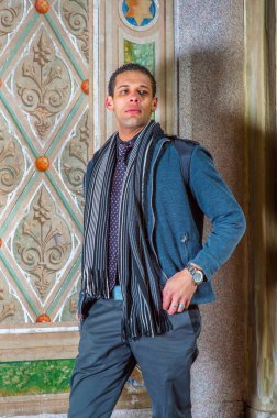 Young man, sitting standing against old wall in winter, thinking, wearing in woolen jacket, tie, long scarf, black pants