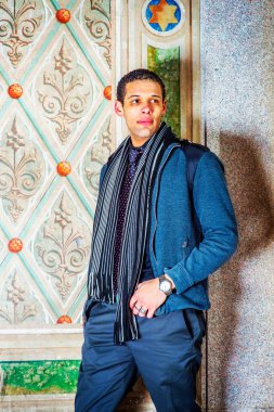Young man, sitting standing against old wall in winter, thinking, wearing in woolen jacket, tie, long scarf, black pants