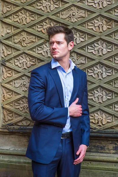 Dressing in a blue slim fit suit and a light blue under shirt, a young handsome businessman is confidently standing there with his hand on heart