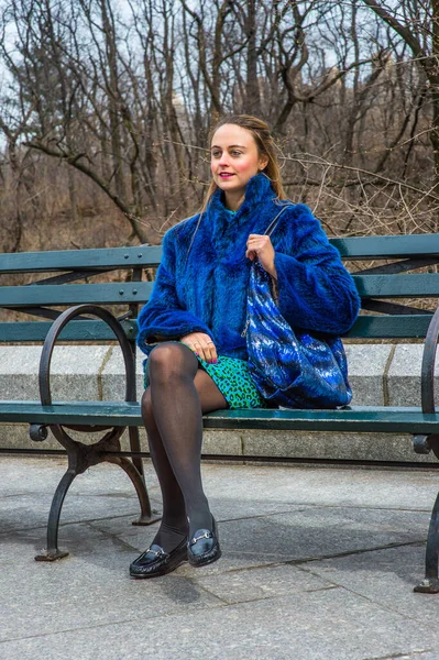青いフェイクファーコートでのドレッシング パターン化されたドレススカート 黒のレギンス 革の靴 バッグを運ぶ 可愛い女の子は 前方を探して 長い椅子に座っている — ストック写真