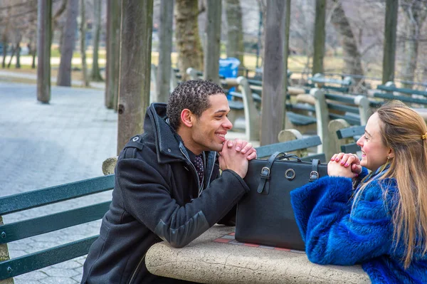 A girl dressing in a blue faux fur plus size jacket and a guy dressing in a black pea coat are sitting crossing a table, smilingly looking each other, with a leather bag