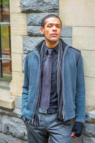 Dressing in woolen jacket with shoulder pads, tie, long scarf and gloves, a young handsome guy is standing against the wall, tired