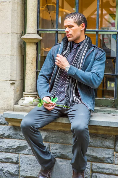 Dressing in woolen jacket with shoulder pads, tie, long scarf, holding a white flower, a young handsome guy with short, curly hair is sitting on the frame of a window, waiting