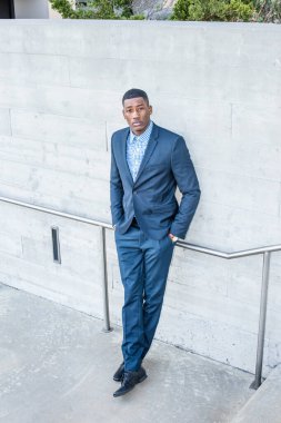 Genç, yakışıklı bir zenci adam dışarıda duvarın önünde duruyor, elleri ceplerinde ve kendinden emin bir şekilde yukarı bakıyor.