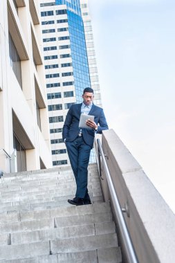 Genç, yakışıklı bir siyahi işadamı bir iş binasının önünde duruyor ve küçük bir bilgisayarın başında kitap okuyor.