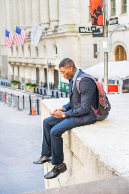 Mavi takım elbise ve deri ayakkabılar giyip sırt çantasıyla ilgilenen genç bir siyahi işadamı dışarıda cep telefonundaki mesajları kontrol ediyor. Arka planda Wall Street tabelası var.