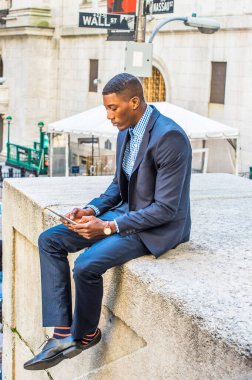 Mavi takım elbise ve deri ayakkabılar giyen genç bir iş adamı dışarıda oturmuş tablet bilgisayarında kitap okuyor.