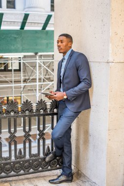 Mavi takım elbise ve deri ayakkabılar giymiş, elinde tablet bir bilgisayar olan genç siyah bir işadamı, dışarıda köşede durmuş, derin düşüncelere dalıyor.