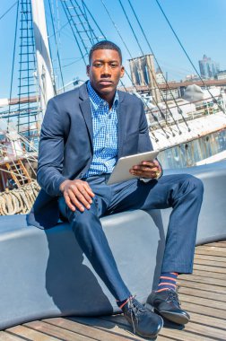 Genç siyah bir işadamı güvertede oturuyor, tablet bilgisayarında kitap okuyor. Arka plan bir liman.