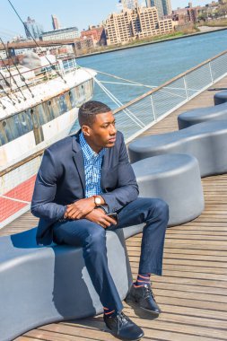 Mavi takım elbiseli, deri ayakkabılı, elinde cep telefonu olan genç bir iş adamı, derin düşüncelere dalmış bir şekilde nehrin kenarında oturuyor.