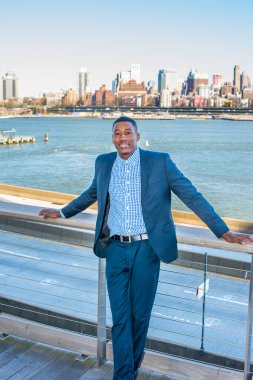 Genç siyah bir işadamı nehir kenarında duruyor, dinleniyor. Arka plan eski bir şehir manzarasıdır.