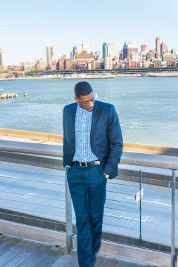 İki elini cebine sokuyor, başını eğiyor, genç siyah bir işadamı derin düşüncelere dalmış nehrin kenarında duruyor. Arka plan eski bir şehir manzarasıdır.