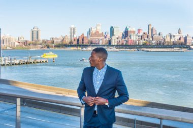 Genç bir siyah işadamı nehir kenarında duruyor, dinleniyor ve düşünüyor. Arka plan eski bir şehir manzarasıdır.