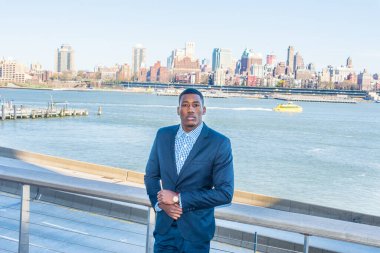 Genç siyah bir işadamı nehrin kenarında duruyor ve kendinden emin bir şekilde sana bakıyor. Arka plan eski bir şehir manzarasıdır.