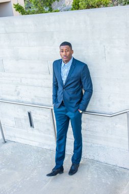 Genç, yakışıklı bir zenci adam dışarıda duvarın önünde duruyor, elleri ceplerinde ve kendinden emin bir şekilde yukarı bakıyor.