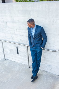 Genç yakışıklı siyah bir adam duvarın önünde duruyor, elleri ceplerinde, bacak bacak üstüne atıyor, uzaklara bakıyor ve seni bekliyor.