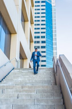 Tablet bilgisayarı tutan, kol saatine bakan genç, yakışıklı bir iş adamı aceleyle bir iş binasının merdivenlerinden aşağı iniyor.