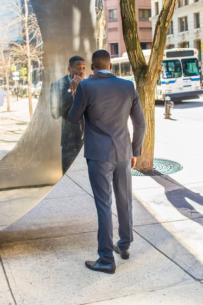 A young black businessman is standing by a mirror wall on the street,  examining himself