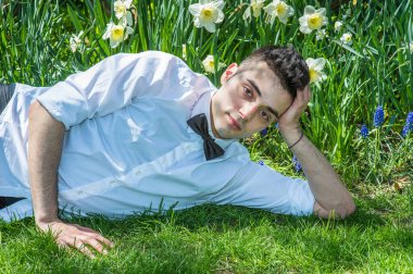 Beyaz gömlek ve siyah papyon takan genç ve yakışıklı bir profesyonel rahatlamak için çiçeklerin önündeki çimlerin üzerinde yatıyor.