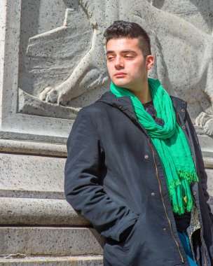 Baharın başlarında güneş ışığı altında, kapüşonlu ceket ve eşarp giymiş, genç yakışıklı bir adam duvarın yanında duruyor, ileriye bakıyor.