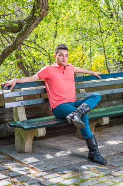 Kırmızı Polo tişörtü, yeşil pantolon ve uzun deri botlar giymişti. Bir kolu başını destekliyordu. Genç ve yakışıklı bir adam bahar sezonunun sabahında uzun bir sandalyede oturuyordu.