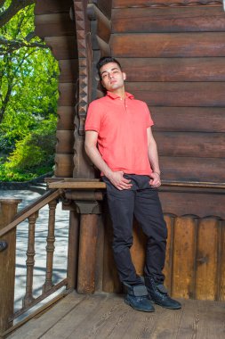 Kırmızı Polo gömlekli, siyah pantolonlu ve botlu, iki eli kalçasında dinlenmiş, genç yakışıklı bir adam eski moda bir tarzda eğiliyor, ahşap ev kapısı, düşünceli bir şekilde sana bakıyor.