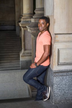 Bir sütuna yaslanmış genç yakışıklı siyah bir adam koridorda duruyor ve sana gülümsüyor.