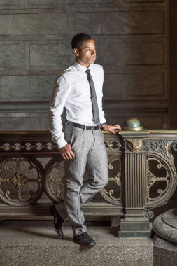 Beyaz gömlekli, siyah kravatlı, gri pantolonlu, genç, yakışıklı bir siyah işadamı korkulukların önünde seni bekliyor.