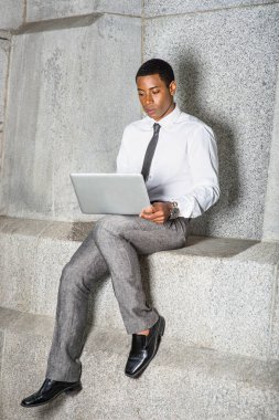 Beyaz gömlekli, siyah kravatlı, gri pantolonlu genç, yakışıklı bir iş adamı dışarıda oturmuş bilgisayar üzerinde çalışıyor.