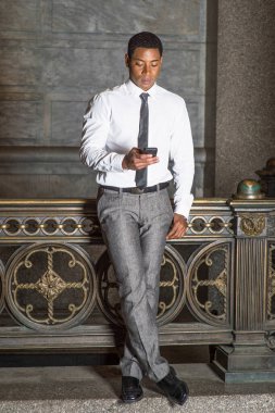 Beyaz gömlekli, siyah kravatlı, gri pantolonlu, genç, yakışıklı bir siyahi işadamı korkuluk başında duruyor ve cep telefonundaki mesajları kontrol ediyor.