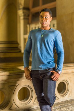 Açık mavi, uzun kollu bir tişört giyen genç, yakışıklı siyah bir öğrenci koridorda duruyor ve kendinden emin bir şekilde ileriye bakıyor.