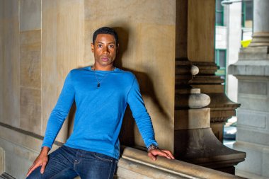 Açık mavi, uzun kollu bir tişört giymiş genç, yakışıklı siyah bir öğrenci bir balkonda duvarın karşısında dikiliyor ve içtenlikle sana bakıyor.