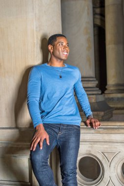 Açık mavi, uzun kollu bir tişört giymiş genç, yakışıklı siyah bir öğrenci koridorda bir duvara yaslanmış gülümsüyor.