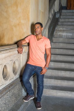Açık turuncu kısa kollu V yaka gömlek, kot pantolon ve deri ayakkabılar giyen genç, yakışıklı bir zenci öğrenci aşağıda dikilip derin düşüncelere dalıyor.