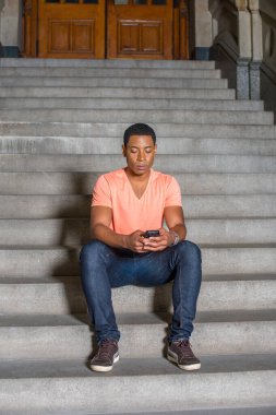 Açık turuncu kısa kollu V yaka gömlek, kot pantolon giymiş genç, yakışıklı bir siyahi öğrenci ofis binasının önünde oturmuş cep telefonundaki mesajları kontrol ediyor..