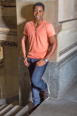 Açık turuncu kısa kollu V yaka gömlek, kot pantolon ve deri ayakkabılar giyen genç, yakışıklı bir zenci öğrenci merdivenlerden duvara dayanıyor ve sana gülümsüyor.