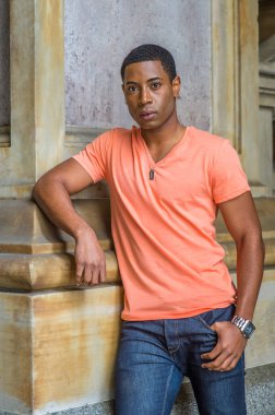Açık turuncu kısa kollu V yaka, kot pantolon giyen yakışıklı bir siyahi öğrenci alt katta bir sütunun yanında duruyor ve kendinden emin bir şekilde sana bakıyor.