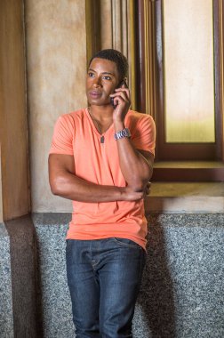 Açık turuncu kısa kollu V yaka gömlek, kot pantolon giyen genç, yakışıklı siyah bir öğrenci küçük bir pencerede durup telefonu dinliyor.
