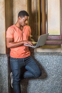Açık turuncu kısa kollu V yaka gömlek, kot pantolon giyen genç, yakışıklı bir siyahi öğrenci küçük bir pencerenin önünde dizüstü bilgisayar üzerinde çalışıyor.
