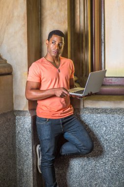 Açık turuncu kısa kollu V yaka, kot pantolon giyen yakışıklı bir siyah öğrenci pencerenin önünde dizüstü bilgisayar üzerinde çalışıyor.