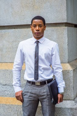 Beyaz gömlekli, siyah kravatlı, gri pantolonlu, evrak çantası taşıyan, taş duvarın yanında duran yakışıklı siyah bir öğrenci, kendine güvenen bir şekilde sana bakıyor..