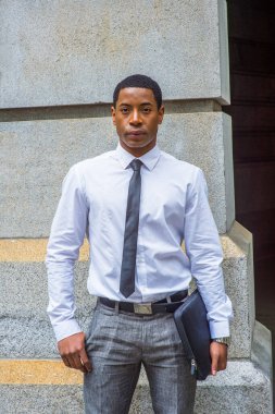 Beyaz gömlekli, siyah kravatlı, gri pantolonlu, evrak çantası taşıyan, taş duvarın yanında duran yakışıklı siyah bir öğrenci, kendine güvenen bir şekilde sana bakıyor..