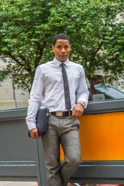Beyaz gömlekli, siyah kravatlı, gri pantolonlu, evrak çantası taşıyan genç, yakışıklı bir siyahi üniversite öğrencisi kampüste durmuş sana gülümsüyor.