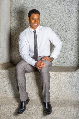 Beyaz gömlekli, siyah kravatlı, gri pantolonlu ve deri ayakkabılı, saat takan, elleri kucağında dinlenen, genç siyah bir öğrenci taş bir bankta oturuyor, gülümsüyor.