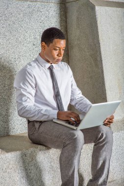 Beyaz gömlekli, siyah kravatlı, gri pantolonlu, genç bir siyahi üniversite öğrencisi ofis binasının dışında bilgisayar üzerinde çalışıyor.