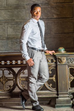 Beyaz gömlekli, siyah kravatlı, gri pantolonlu, üniversiteli siyahi bir erkek öğrenci kampüste korkulukların önünde duruyor ve kendinden emin bir şekilde sana bakıyor.