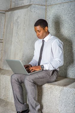 Beyaz gömlekli, siyah kravatlı, gri pantolonlu, genç bir siyahi üniversite öğrencisi ofis binasının dışında bilgisayar üzerinde çalışıyor.