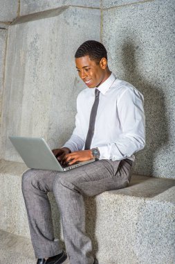 Beyaz gömlekli, siyah kravatlı, gri pantolonlu, genç bir siyahi üniversite öğrencisi ofis binasının dışında bilgisayar üzerinde çalışıyor..