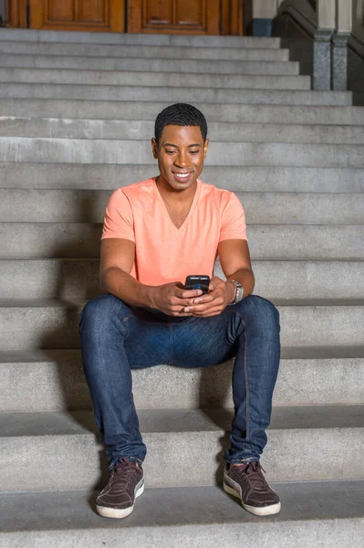 Dressing in a light orange short sleeve V neck shirt, jeans, a young handsome black student is sitting on stairs outside an office building, checking messages on his mobile phone.